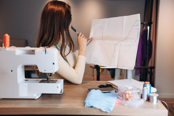 Young dressmaker woman sews clothes on sewing machine and creating new project in workshop. Creating online clothing design courses. - 写真・画像