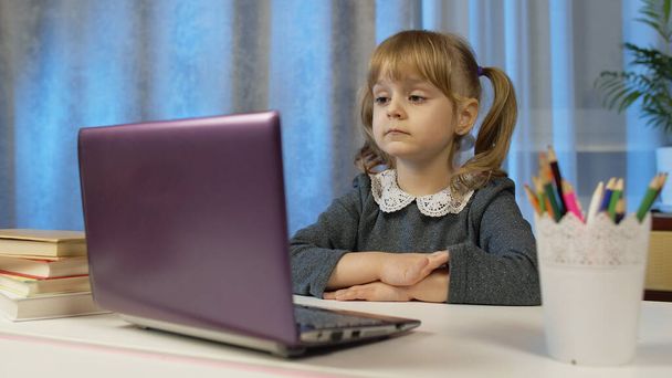 Lapsi tyttö tekee koulun kotitehtäviä kirjoittaa muistikirjaan, käyttäen kannettavaa tietokonetta kotona, online-koulutus - Valokuva, kuva