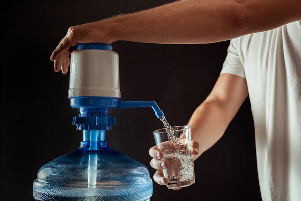 男はハンドポンプを使ってコップに飲料水を注ぐ。黒の背景に純水の大きなボトル - 写真・画像