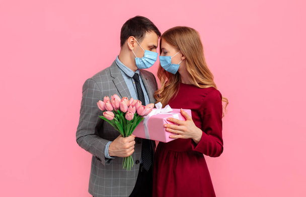 Valentine's day celebration, happy couple in love, man and woman wearing medical masks on face, to protect from virus, with gift and bouquet of flowers on isolated pink background - Fotoğraf, Görsel