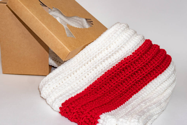 В'язаний шарф біло-червоно-білий акуратно складений поруч з картонною коробкою на білому тлі крупним планом
 - Фото, зображення