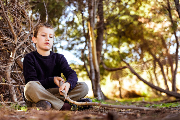 A child sitting on the ground in a forest uses a stick to draw on the ground and experience nature. - Photo, Image