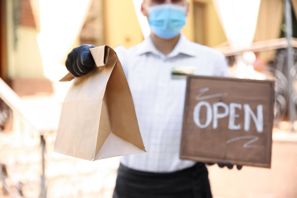 Waiter with packed takeout order and OPEN sign near restaurant, closeup. Food service during coronavirus quarantine - Zdjęcie, obraz