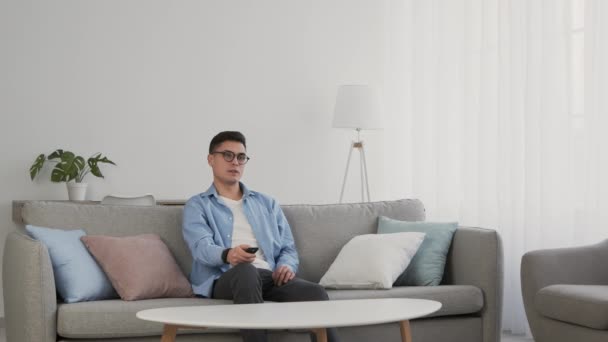 Young boring man in glasses switching channels on tv with remote controller and feeling irritated, slow motion - Footage, Video