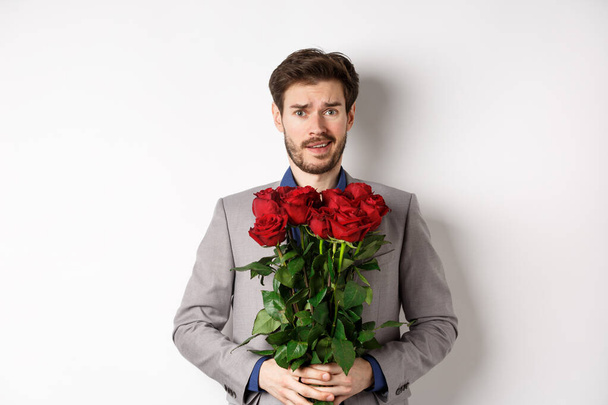 ustaraný přítel v obleku, držící květiny růže a vypadající pochybovačně na kameru, stojící s kyticí na Valentýna na bílém pozadí - Fotografie, Obrázek