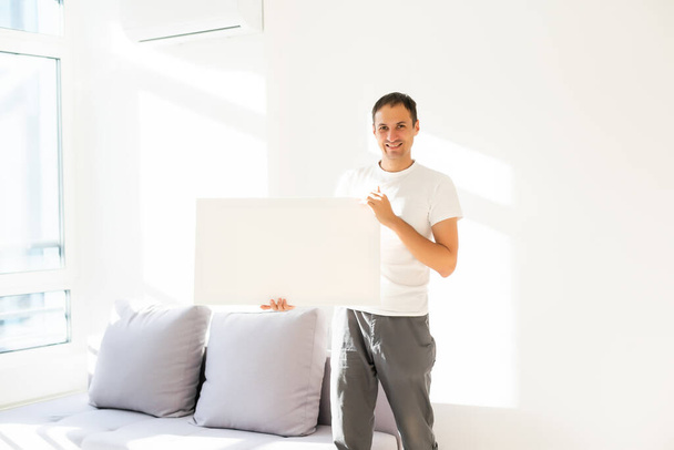 Un jeune homme en t-shirt blanc tient une toile vide. Un étudiant sourit. Modèle pour la publicité, salutation, invitation. Portrait émotionnel - Photo, image