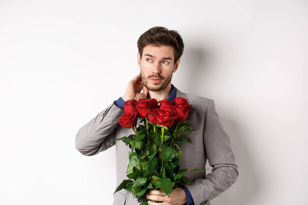 Pensive young man in suit holding bouquet of flowers, waiting for date on Valentines day, standing over white background - Фото, зображення