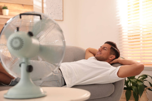 Man enjoying air flow from fan on sofa in living room. Summer heat - Фото, изображение