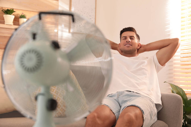 Man enjoying air flow from fan on sofa in living room. Summer heat - Fotoğraf, Görsel