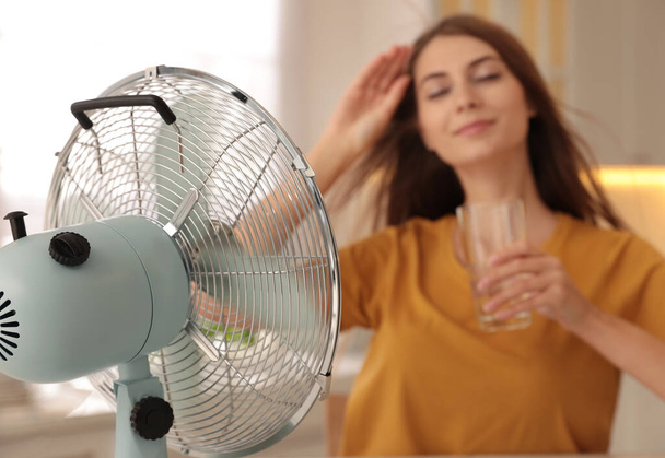 Woman with glass of water in kitchen, focus on fan. Summer heat - Photo, image