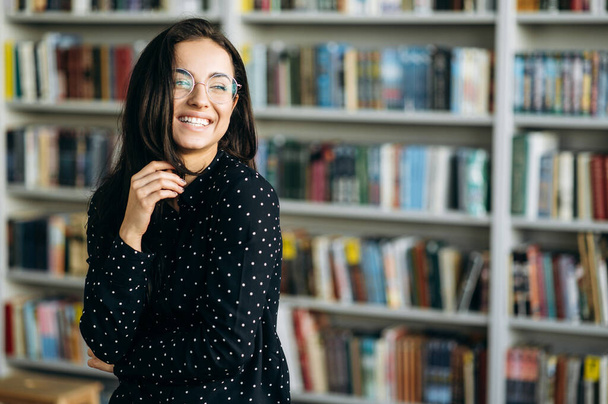 Iloinen tyylikäs valkoihoinen nainen freelancer tai opiskelija, silmälasit ja muodollinen kuluminen seisoo modernissa toimistossa tai yliopiston kirjastossa näyttää pois ja hymyilee - Valokuva, kuva