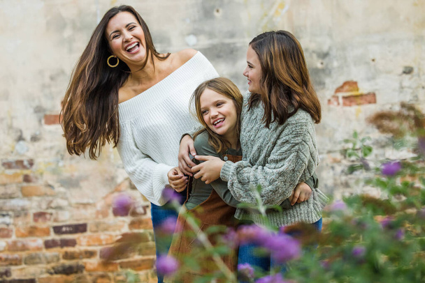 Молодая мама лет тридцати с двумя дочерьми на улице обнимаются и смеются вместе - Фото, изображение