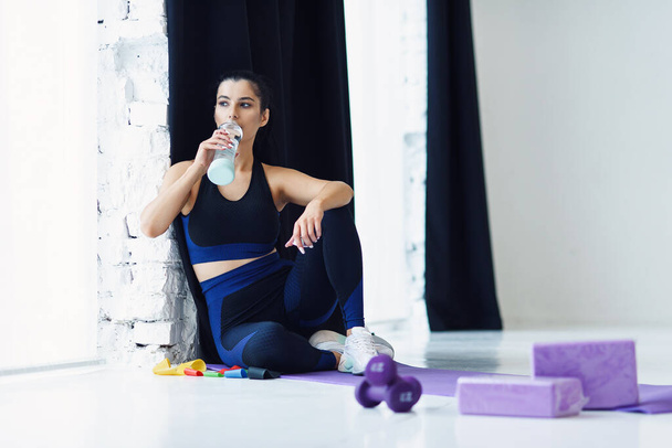Strong women in sportswear sitting on yoga mat and drinking water after workout at home. Health care concept. - Фото, изображение