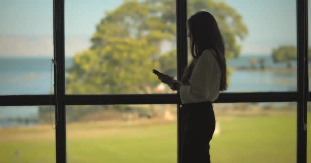 Młoda bizneswoman sprawdzająca telefon stojąc przy dużym oknie i patrząc na zewnątrz. Wolny ruch, średni strzał. - Materiał filmowy, wideo