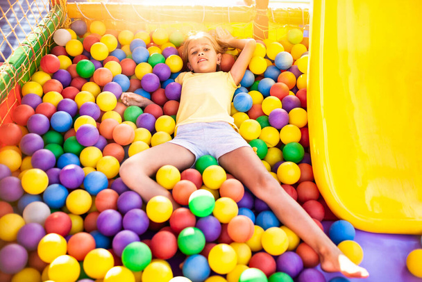 A cute cheerful girl lies on the playground with soft and bright equipment and throws colorful balls into the camera - Photo, Image