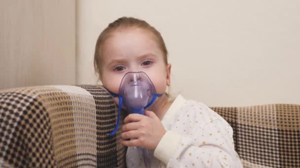 A cute little baby in a mask breathes through an inhaler. Home inhalation procedure. The child is receiving respiratory therapy with a nebulizer. Treat kid from coronavirus at home. A little girl - Footage, Video