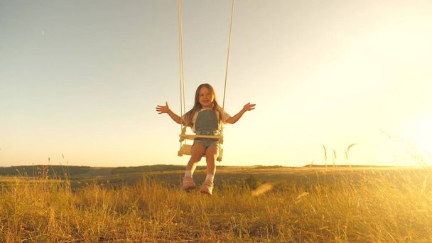 Onnellinen lapsi lentää keinussa puistossa. Pieni iloinen tyttö nauraa, unelmoi lentäjäksi tulemisesta ja lentää kuin lentokone. Äiti ravistelee tervettä tytärtään keinussa puun alla auringossa. Lapsuus ja perhe. - Valokuva, kuva