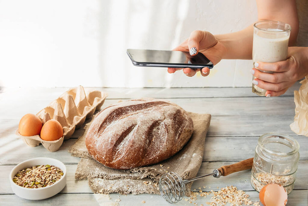 unrecognizable person takes pictures of cooking homemade food on smart phone. homemade baked grain bread with seeds. kitchen table with eggs, flour and pastries. food blogging, online cooking at home - Foto, Bild