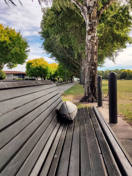 Piłka rugby na ławce w parku publicznym bez ludzi w pobliżu. Fotografia pionowa - Zdjęcie, obraz