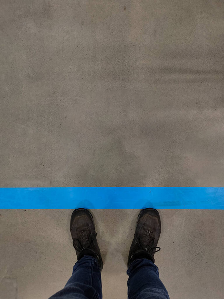Vista superior de los zapatos de hombre de pie en la línea azul de una cola en el mercado para mantener la distancia social. Fotografía vertical - Foto, Imagen