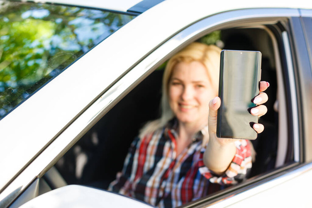 Mooie vrouw zit in een auto en toont haar smartphone uit het raam - Foto, afbeelding
