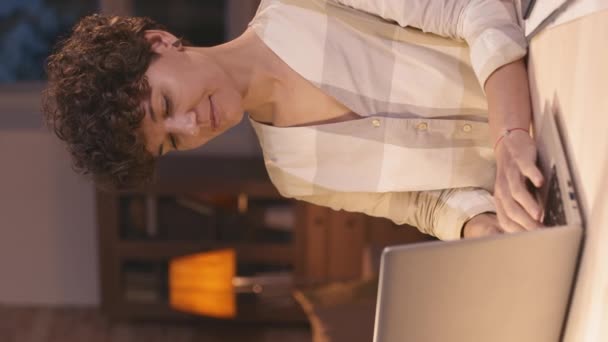 Vertikales Porträt einer fröhlichen jungen Frau, die zu Hause am Schreibtisch sitzt und am Laptop arbeitet und dann für die Kamera lächelt - Filmmaterial, Video