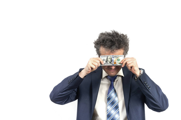 Ongeschoren man van middelbare leeftijd in een zakenpak met slordig haar glimlachend verbergt zijn ogen achter een biljet van 100 dollar op een witte achtergrond. - Foto, afbeelding