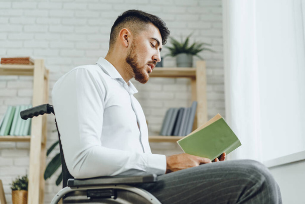 Homem com deficiência em cadeira de rodas lendo um livro dentro de casa - Foto, Imagem