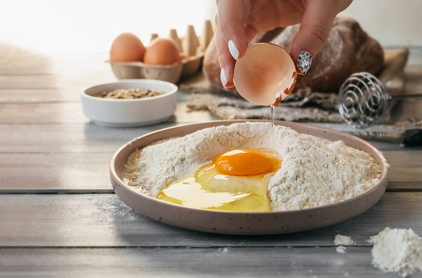 Rohes Ei in der Hand wird gebrochen und in eine stilvolle Schüssel mit Mehl auf dem Hintergrund der Zutaten für das Backen zu Hause gegossen. Selektiver Fokus - Foto, Bild