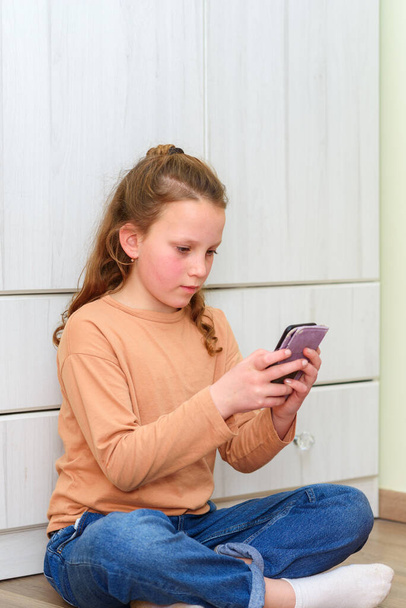 Ragazza seduta sul pavimento e sms sullo smartphone in una casa con un armadio sullo sfondo. - Foto, immagini