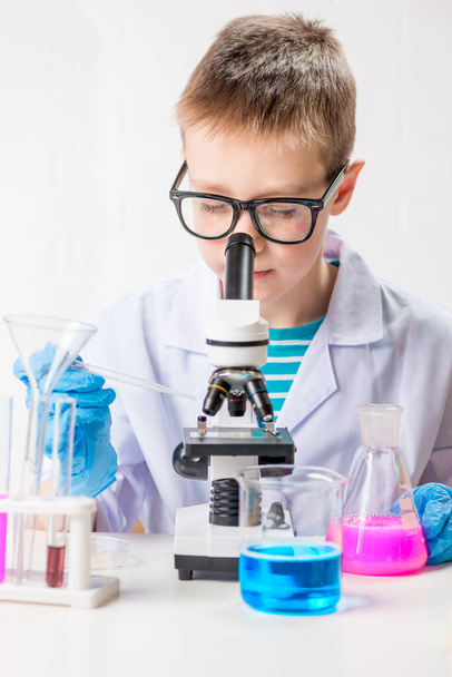 A schoolboy with a microscope examines chemicals in test tubes, conducts experiments - a portrait on a white background. Concept for the study of coronavirus in the laboratory - Foto, imagen