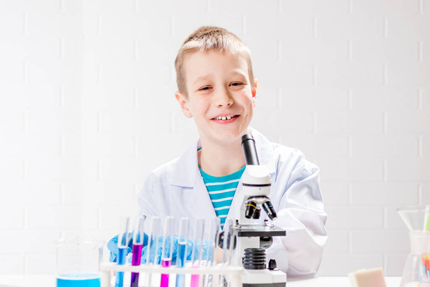 Mikroskoopilla varustettu koulupoika tutkii kemikaaleja koeputkessa, tekee kokeita - muotokuva valkoisella pohjalla. Konsepti koronaviruksen tutkimiseksi laboratoriossa - Valokuva, kuva
