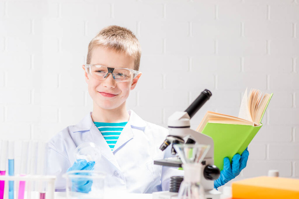 Школьник с микроскопом и книгой исследует химические вещества в пробирках, проводит эксперименты - портрет на белом фоне. Концепция исследования коронавируса в лаборатории - Фото, изображение