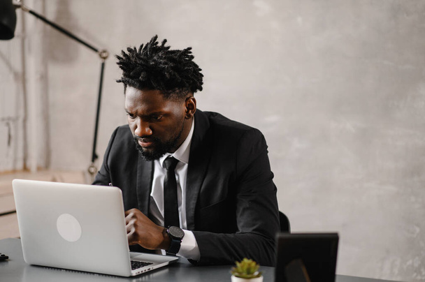  Handsome african american businessman in classic suit uses laptop and smiles while working in office - Foto, afbeelding