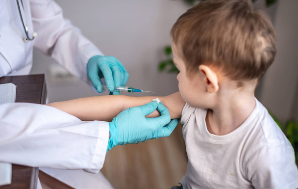 A young female doctor in a white medical coat, blue gloves, and a protective mask is vaccinating a small boy patient in his arm. Concept of health care and vaccination. - Photo, image