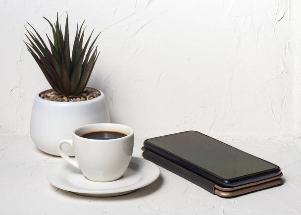 taza blanca con café aromático negro sobre un fondo blanco abstracto con una flor en maceta en el fondo con un teléfono inteligente. - Foto, Imagen