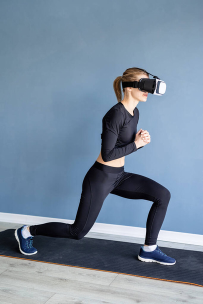 Fitness, sport and technology. Young athletic woman wearing virtual reality glasses doing front lunge working out at home at blue background - Фото, изображение