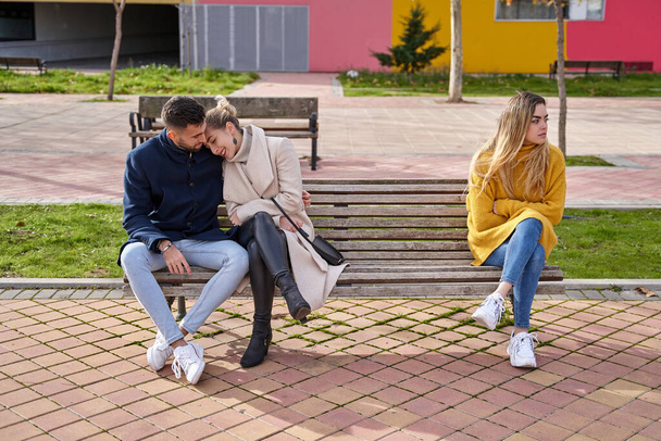 Una pareja comprometida sentada en un banco abrazándose mientras un amigo está solo. Concepto de pareja y angustia - Foto, imagen