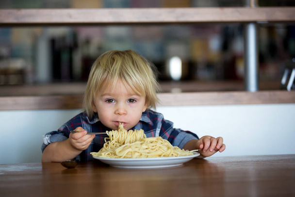 Little baby boy, peuter kind, eten spaghetti als lunch en maken het voeden teddybeer vriend, pot met spaghetti en tomaten op de tafel - Foto, afbeelding