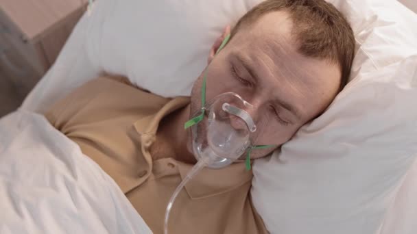 Top view keskipitkän lähikuva nuori valkoihoinen mies yllään happinaamari, makaa sairaalassa sängyssä, avaa silmänsä ja katselee kameraan - Materiaali, video