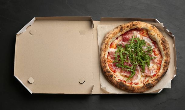 Tasty pizza with meat and arugula in cardboard box on black table, top view - Foto, afbeelding