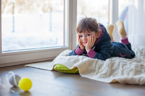 Adorable little girl lying on a woolen blanket near the window, it's snowy winter outside - Foto, afbeelding