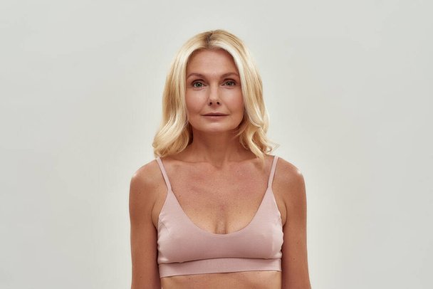 Human age. Portrait of a beautiful middle aged blonde woman in pink bra looking at camera while posing isolated over grey background - Foto, immagini