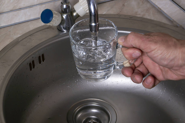 мужчина держит стакан кружки под струей чистой питьевой воды - Фото, изображение