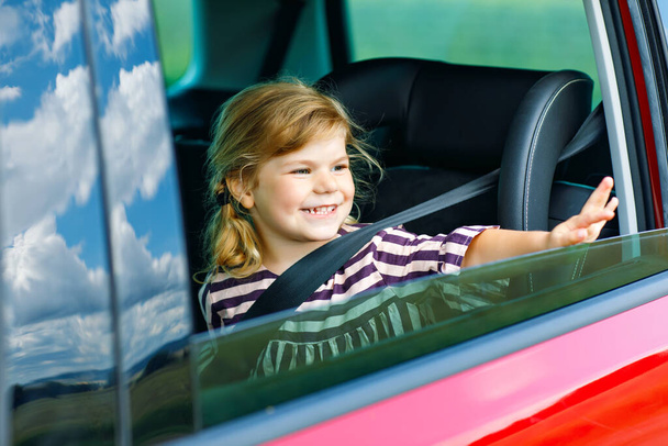 Urocza dziewczynka siedząca w foteliku samochodowym i wyglądająca przez okno na przyrodę i korki. Mały dzieciak podróżujący samochodem. Bezpieczeństwo dzieci na drodze. Wycieczka rodzinna i wakacje w lecie - Zdjęcie, obraz