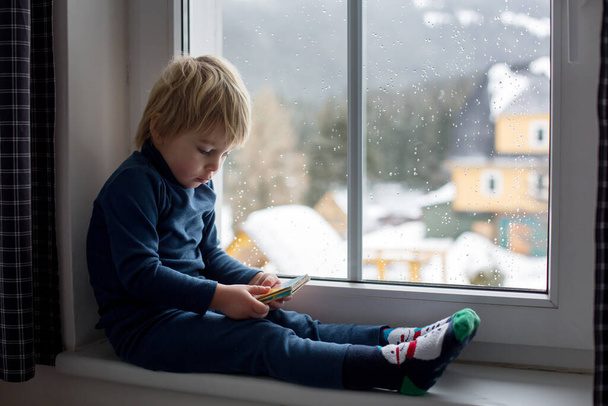 Toddler child, sitting on the window, watching the snow falling, reading little book - Фото, изображение
