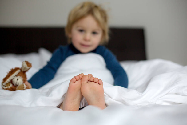 Little toddler child feet on the foreground, cute child, lying in bed, playing with little stuff toy, smiling happily - Fotoğraf, Görsel
