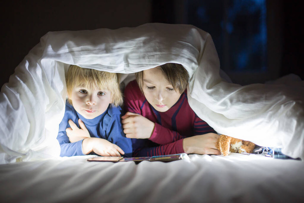Очаровательный блондин ребенок и старший брат, милые мальчики, чтение книжечки с маленькой игрушкой рядом с ним под одеялом в постели - Фото, изображение