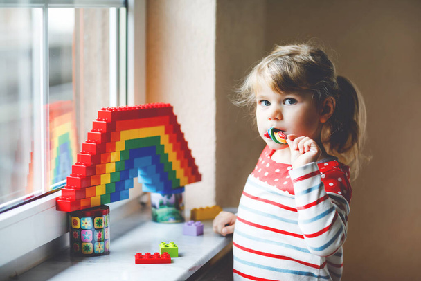 Cute little toddler girl by window create rainbow with colorful plastic blocks during pandemic coronavirus quarantine. Children made and paint rainbows around the world as sign. Kid eating lollipop. - Photo, Image