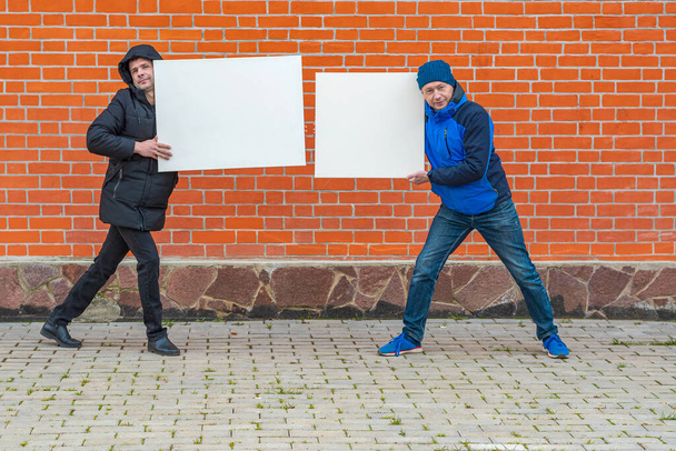 Dos hombres vestidos de negro y azul se paran sobre el fondo de una pared de ladrillo rojo y sostienen en sus manos lienzos blancos en blanco para pintar - Foto, imagen